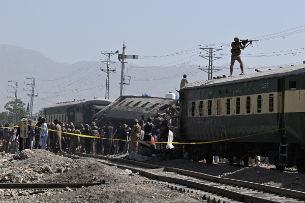 2026年5月24日，巴基斯坦奎達（Quetta），一列列車遭爆炸襲擊後脫軌，一名巴基斯坦士兵站在車廂頂部持槍警戒，安全人員正在現場檢查受損車廂。